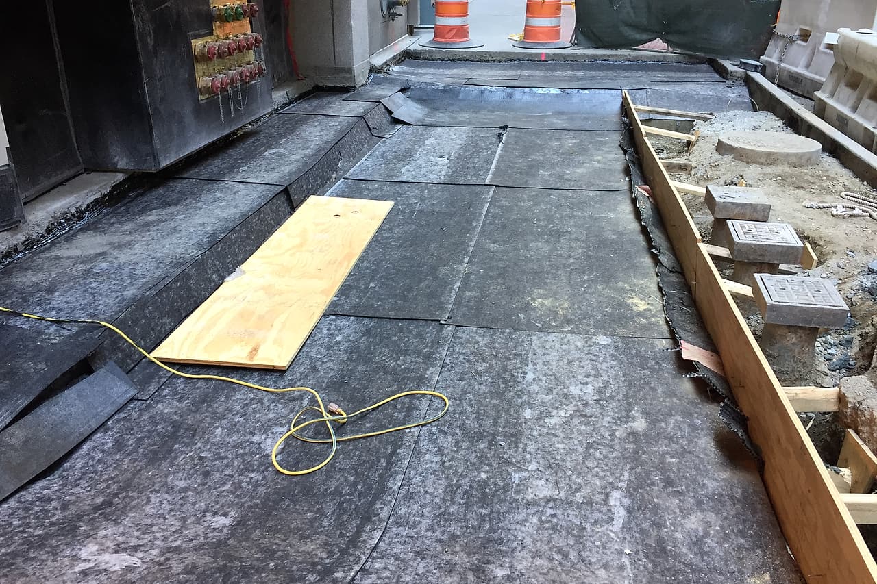 Dark membrane sheets cover an unfinished exterior floor beside a concrete wall, with laps and temporary protection visible.