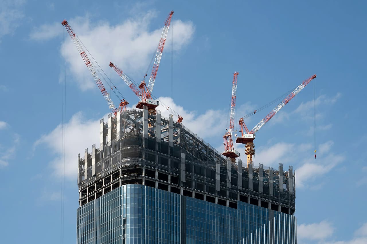 Five tower cranes stand on top of a glass high-rise as its structure nears the top.