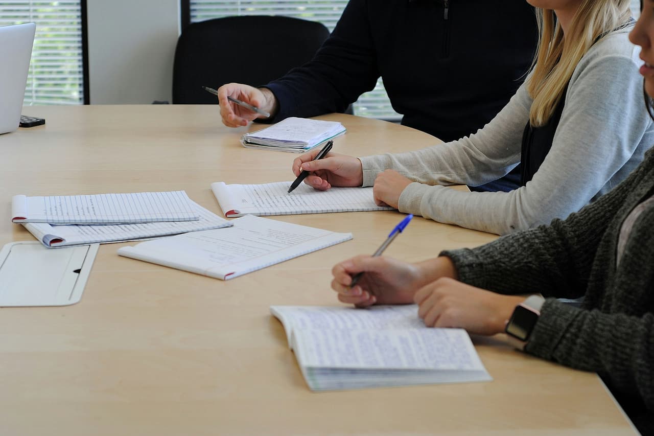 Three people at a meeting-room table take notes by hand on notepads, with more pages of notes spread across the table.
