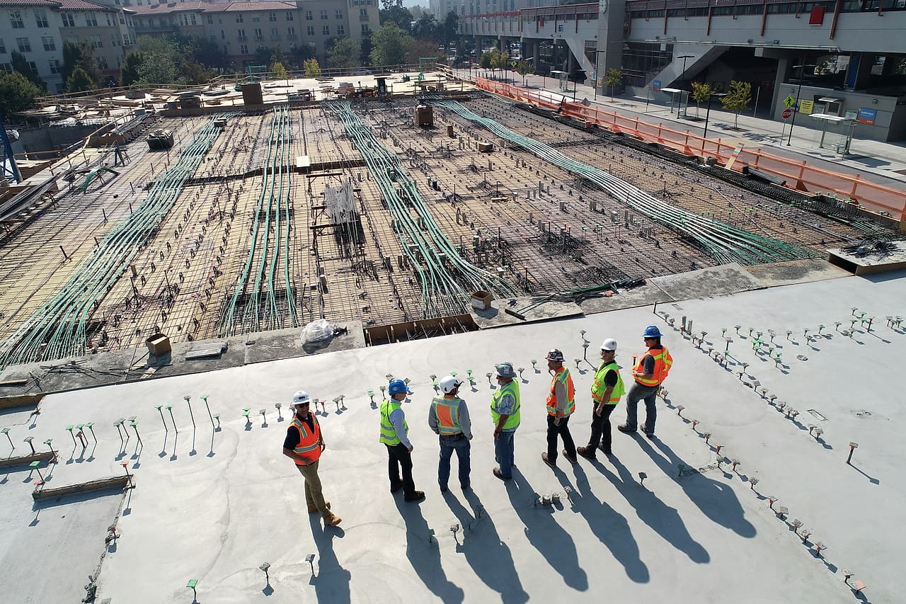 Seven workers in hard hats and high-visibility vests talk in a loose semicircle on a concrete deck beside rebar laid for the next pour.
