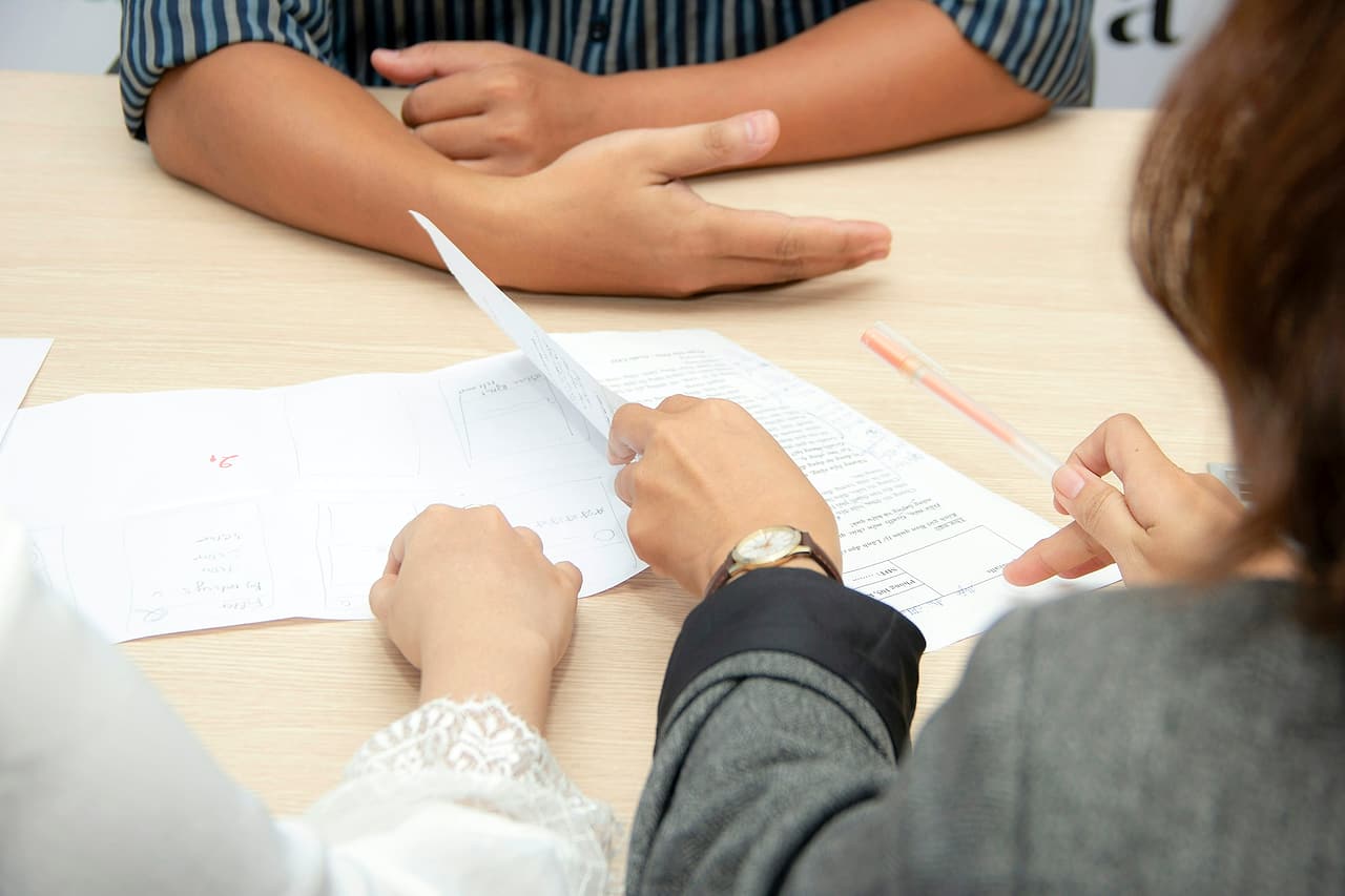 Across a meeting table, one person gestures with an open hand while two others hold a marked-up document and a pen.