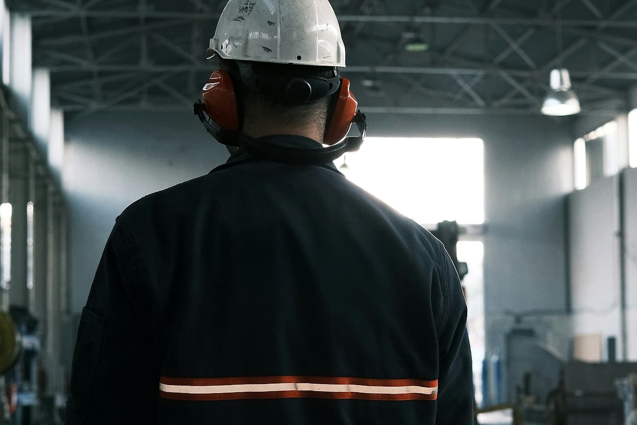A worker wearing a hard hat and hearing protection stands inside an industrial workspace.