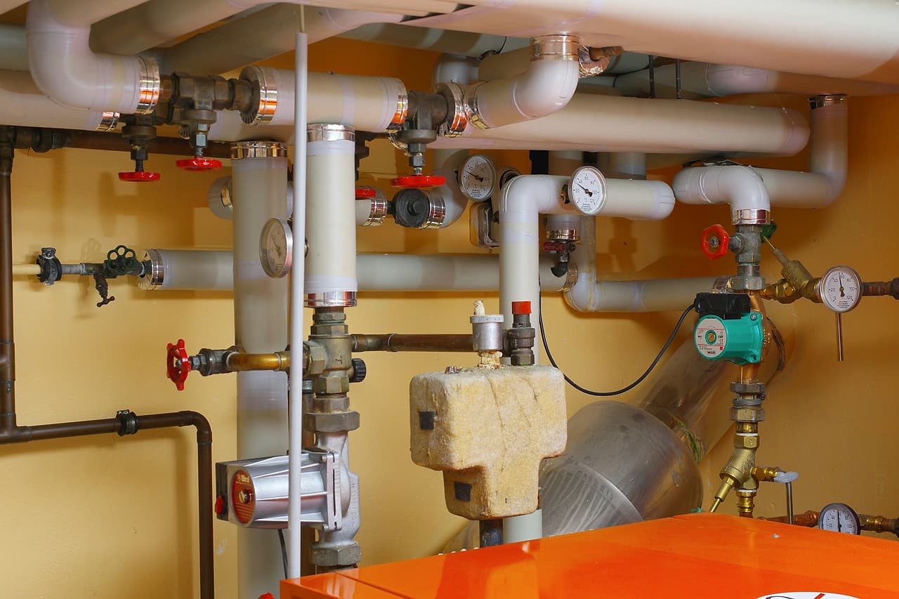 A small boiler room with insulated pipes, red valve handles, temperature gauges and a circulating pump above an orange boiler.