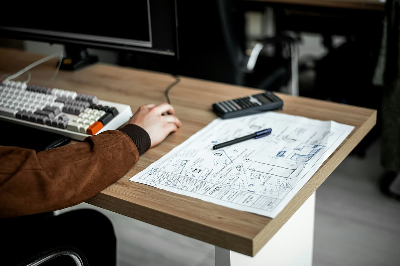A person at a desk works beside a sketched plan sheet, with a pen and a calculator next to the keyboard.