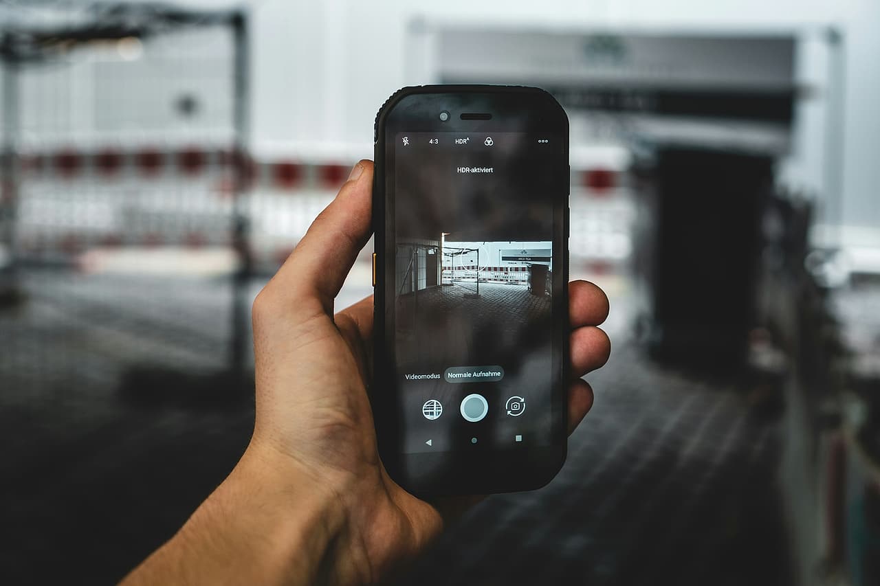 A hand holds up a phone with its camera open, framing a construction site behind red-and-white barriers.