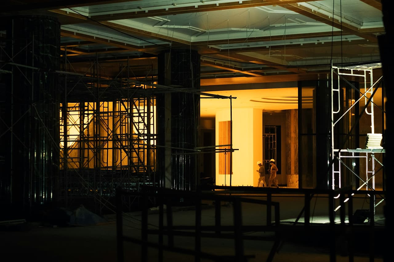 A building interior under construction lit up at night, with scaffold towers and two workers in hard hats walking through.