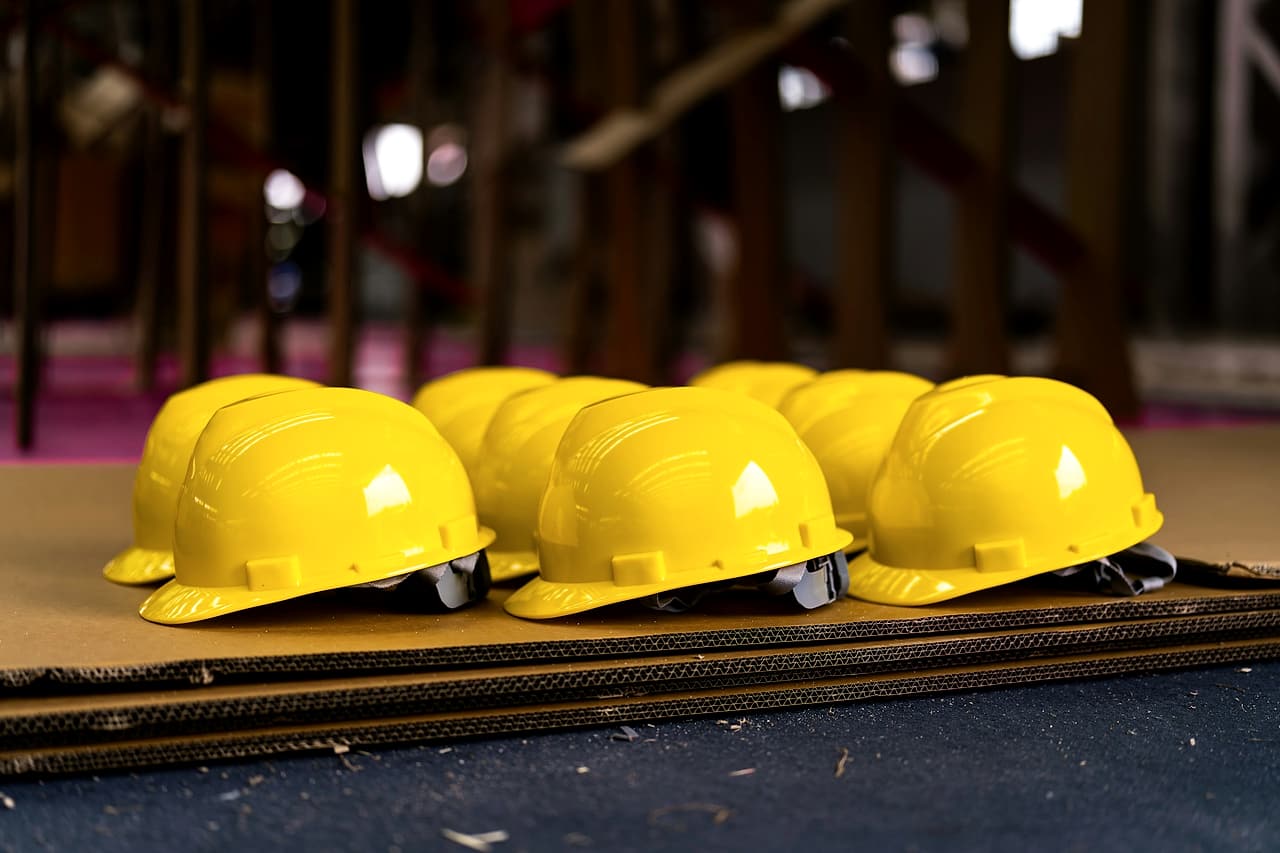 A row of new yellow hard hats laid out on sheets of cardboard, with a building interior blurred behind.
