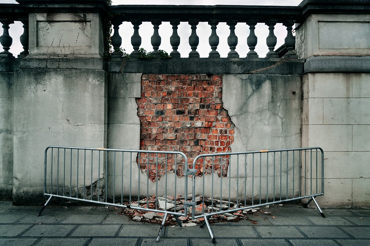 A damaged wall with its facing broken away to the brick, fenced off on the sidewalk with crowd barriers around the debris.