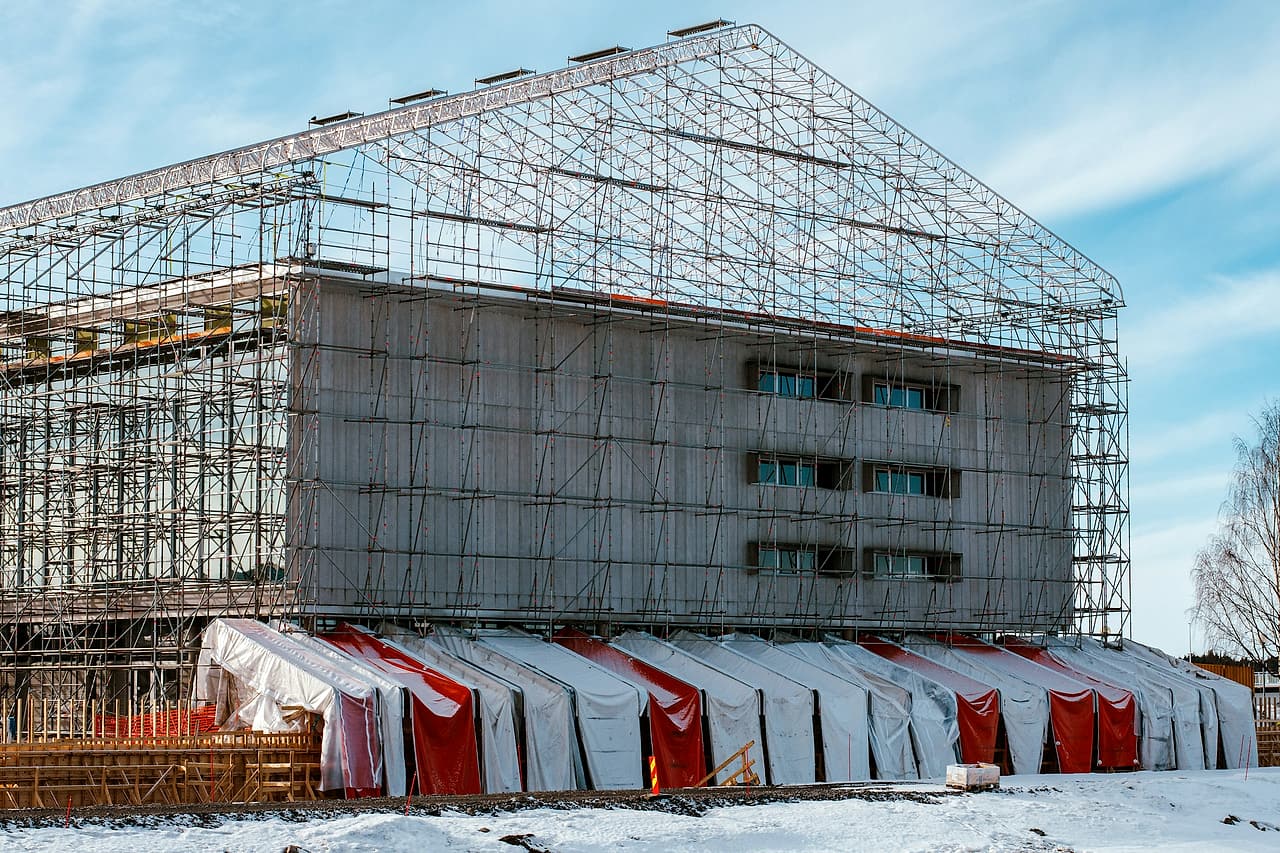 A building under construction sits inside a full scaffold with a temporary roof, its base wrapped in tarps, on snowy ground.