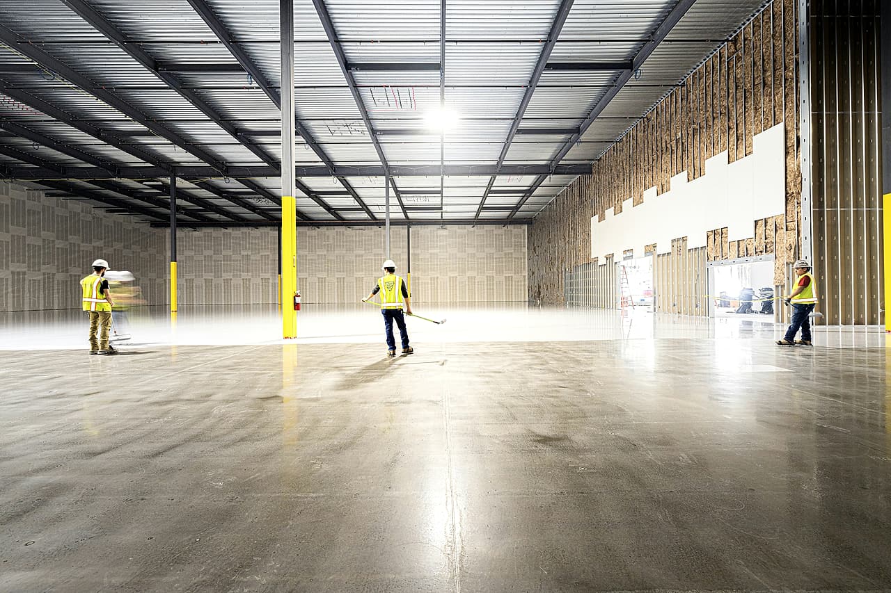 Workers in hard hats and vests apply a floor finish in a nearly complete warehouse while drywall goes up on the far wall.