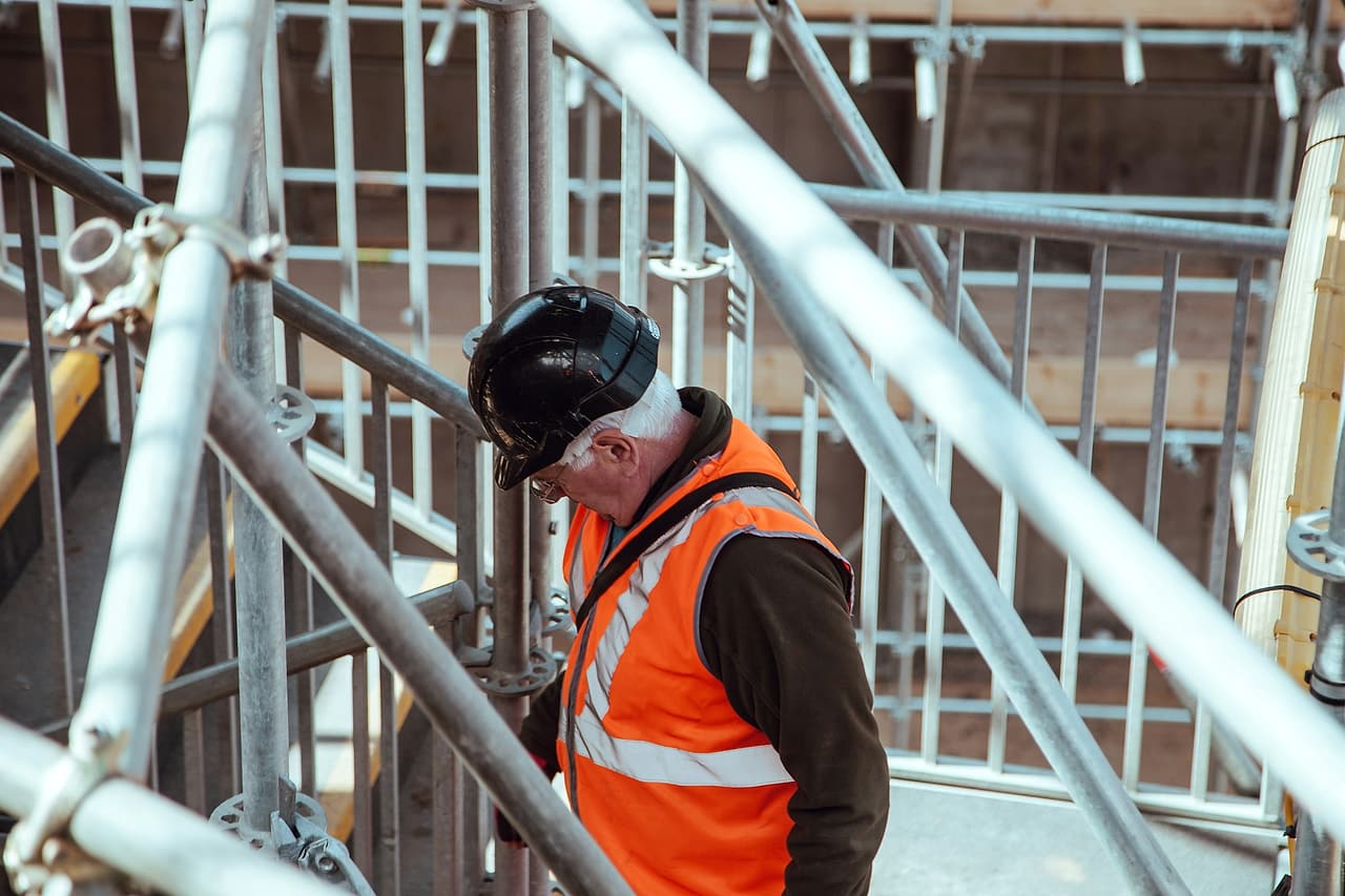 A site supervisor in an orange high-visibility vest and black hard hat on a scaffold stair tower with guardrails.