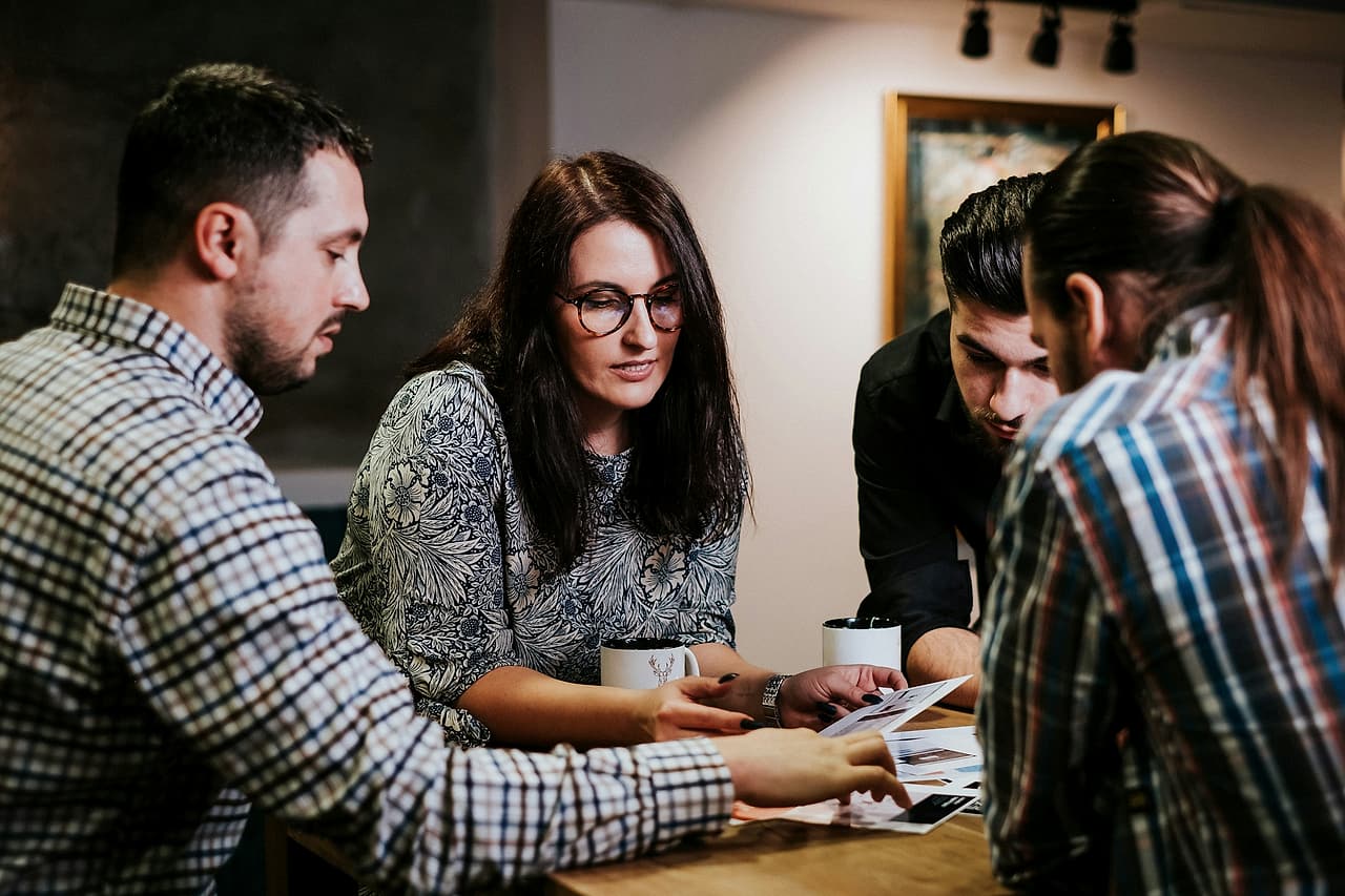 Four people lean over printed sheets on a wooden table, reviewing them together.