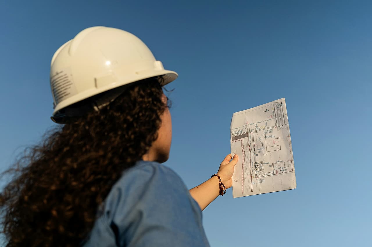 A woman in a white hard hat, seen from behind, holds up a marked-up site plan against a clear blue sky in late-afternoon light.