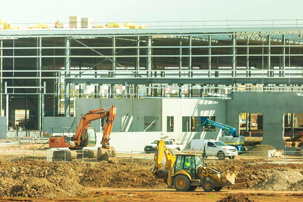 A busy site with an excavator, a backhoe loader, a boom lift and trucks working in front of a precast and steel-framed building.