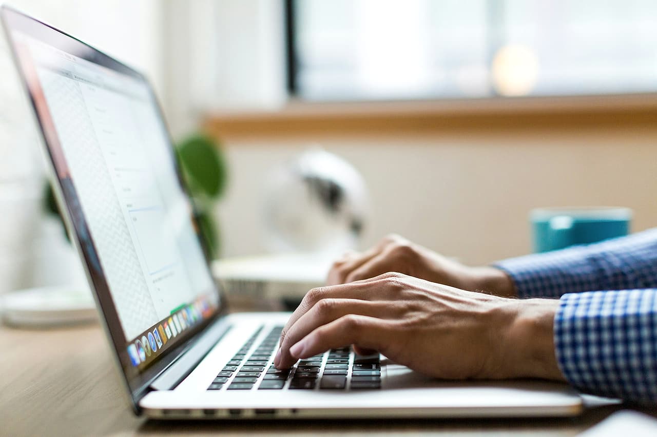 Hands in a checked shirt type on a laptop at a desk by a window, a mail list open on the screen.