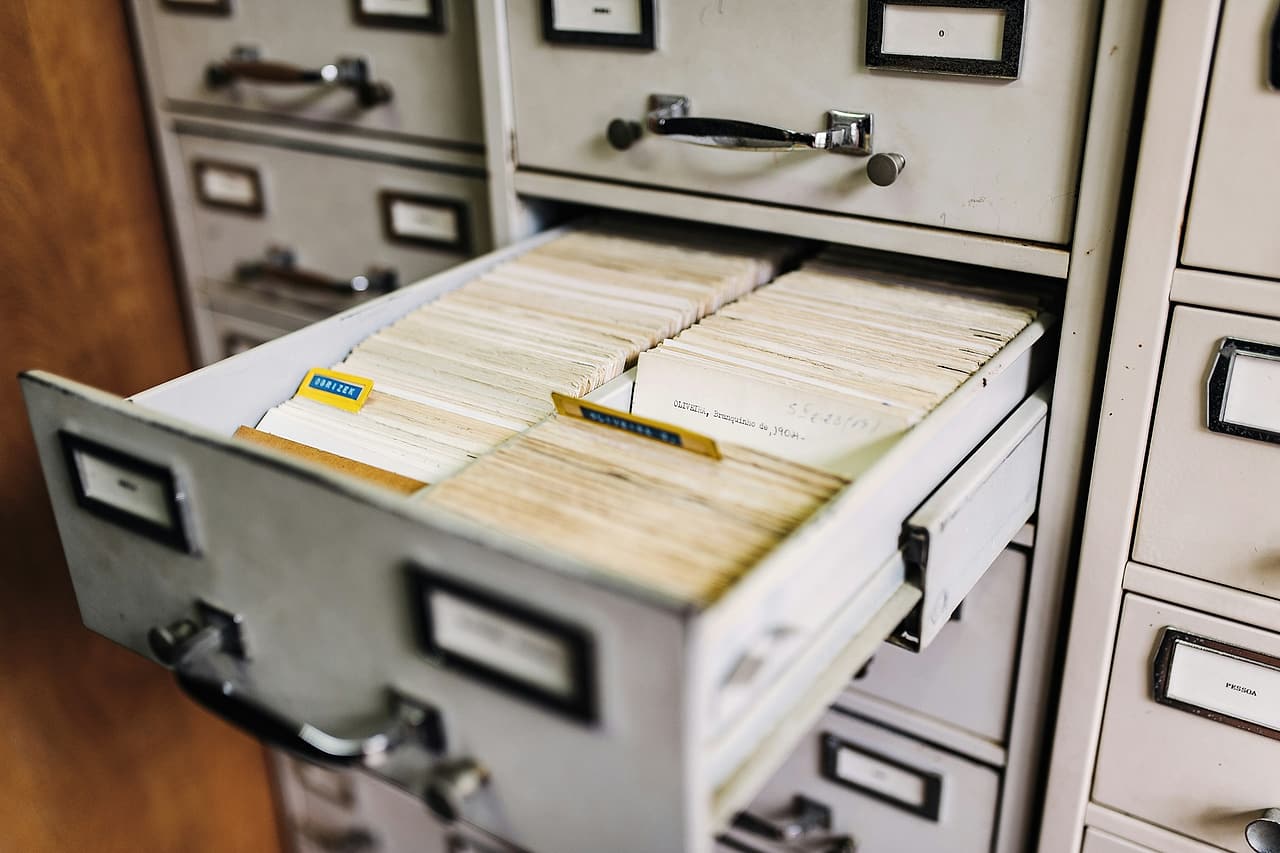 An open drawer of a card-index cabinet, its cards divided by yellow and blue tabs, among rows of labelled drawers.