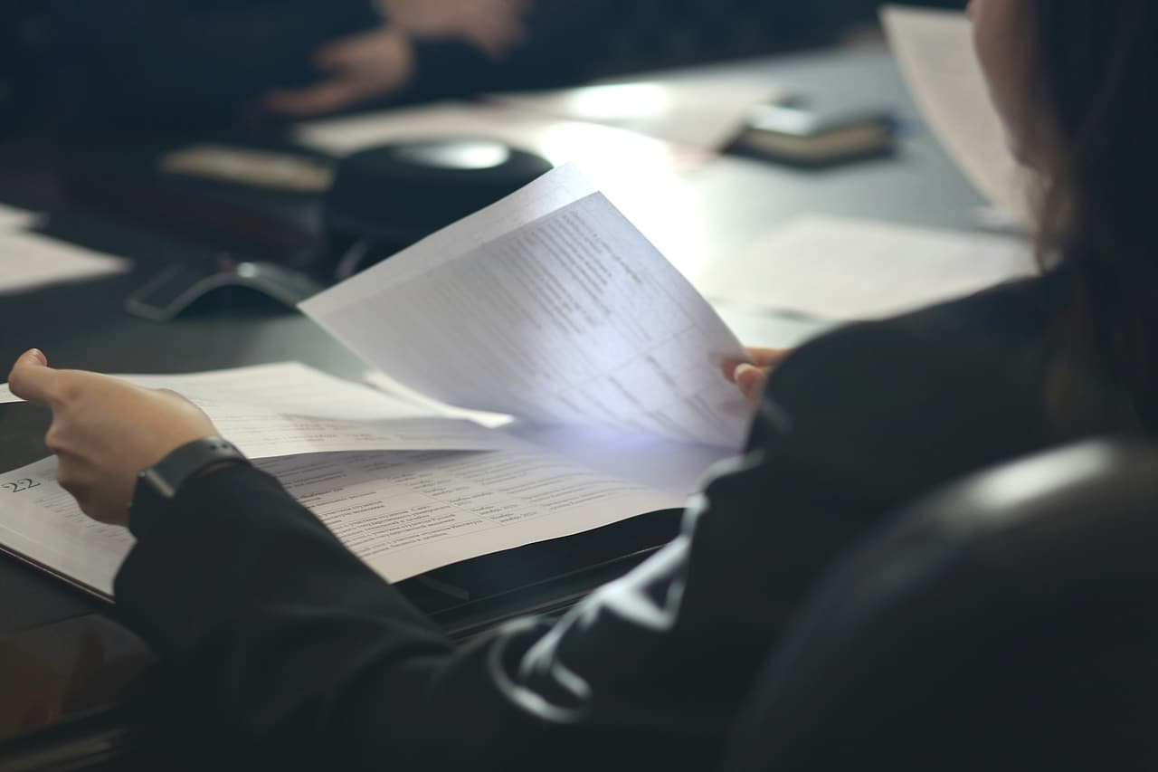 Hands hold a printed document open at a meeting table while other papers and participants blur in the background.