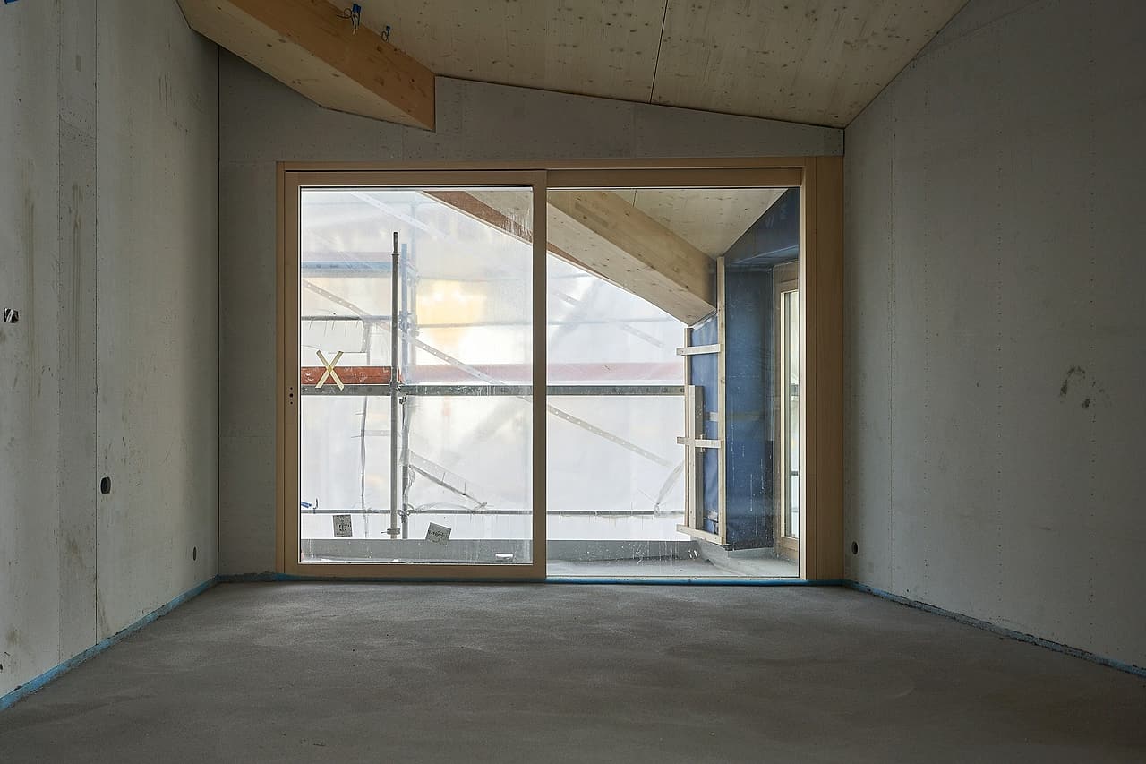 An empty unfinished room has a sliding door and scaffolding visible outside.
