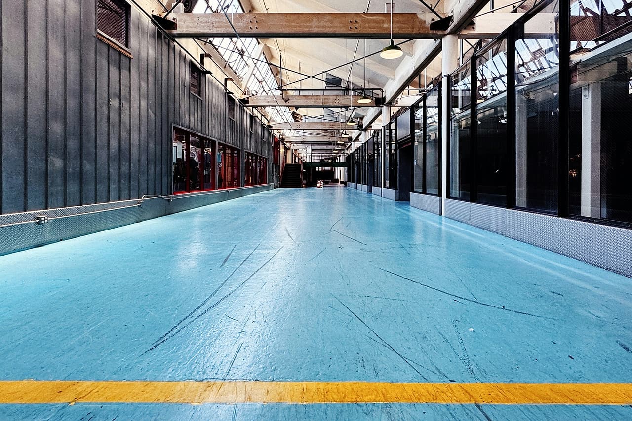 A long corridor in a converted industrial building, its floor covered wall to wall in blue temporary protection with a yellow line.