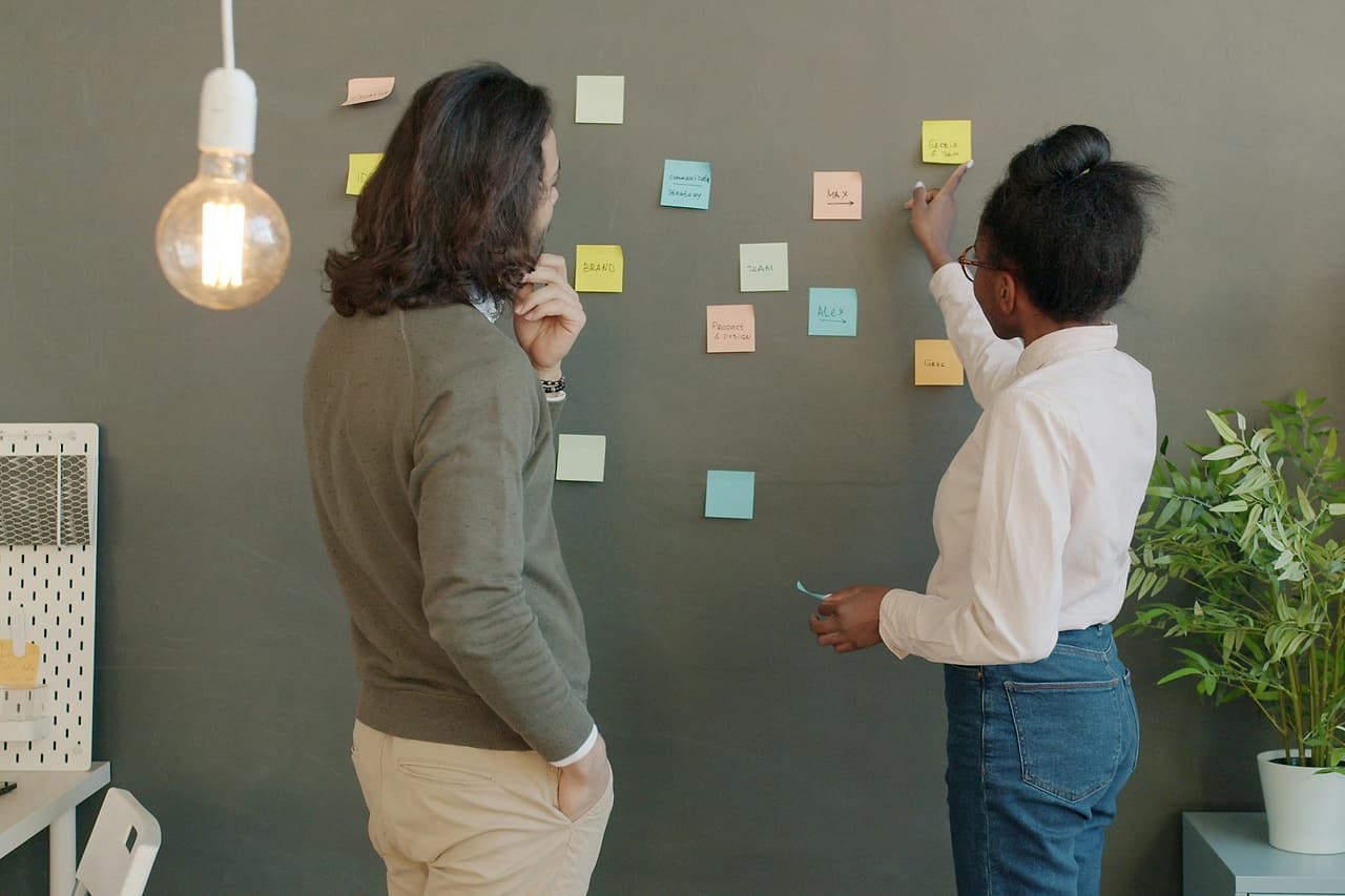 Two colleagues place and study sticky notes on a grey meeting-room wall.