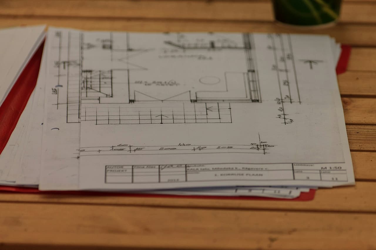 A stack of printed floor plans and a red folder on a wooden table.