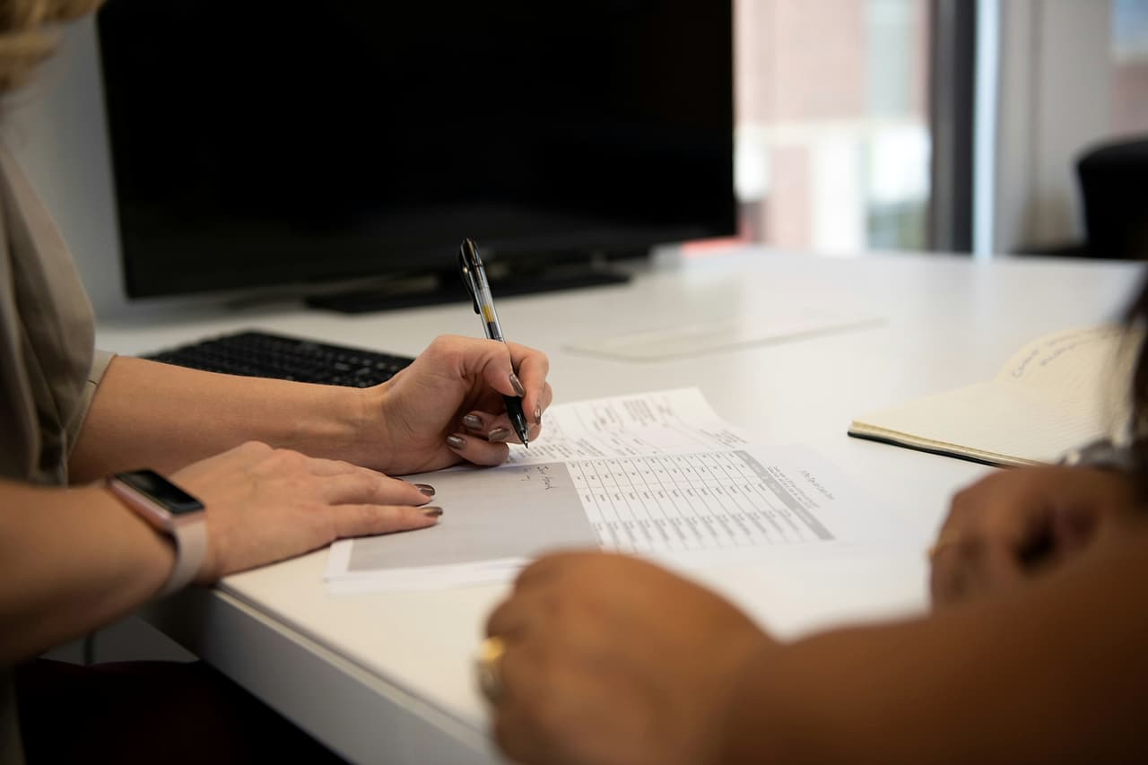 One person marks figures on a printed spreadsheet with a pen while a colleague sits across the desk.