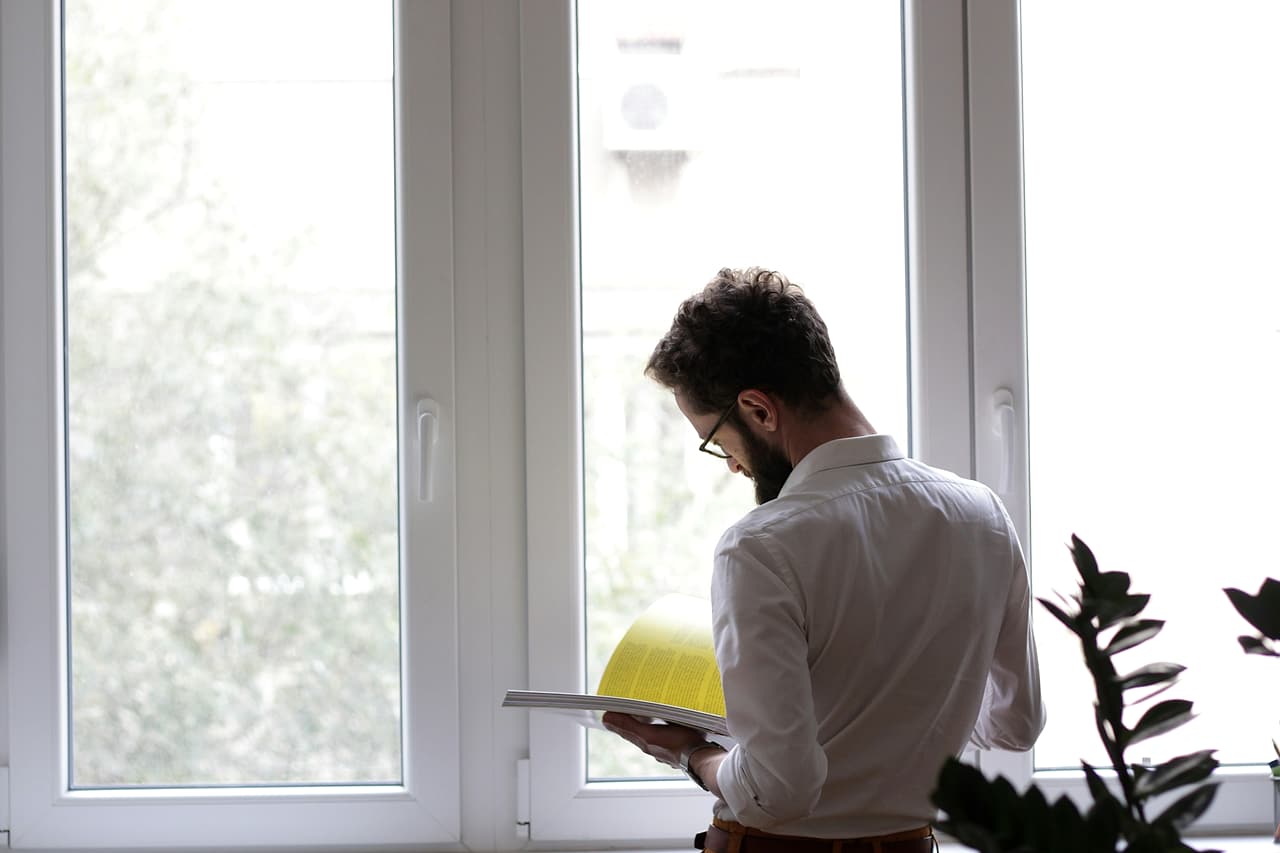A man in a white shirt stands at a bright window reading a thick document with yellow pages, seen from behind.