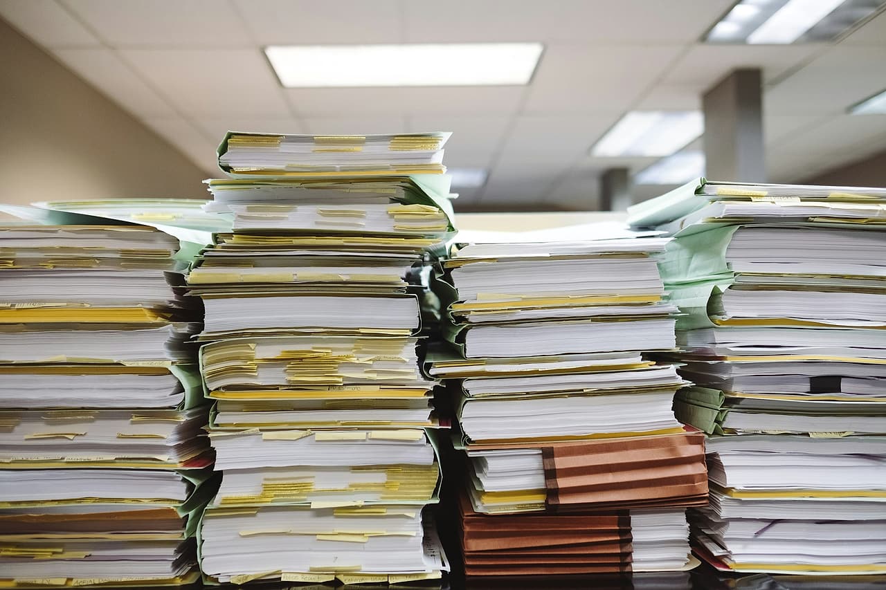 Tall stacks of paper documents and file folders, flagged with yellow sticky tabs, fill an office table.