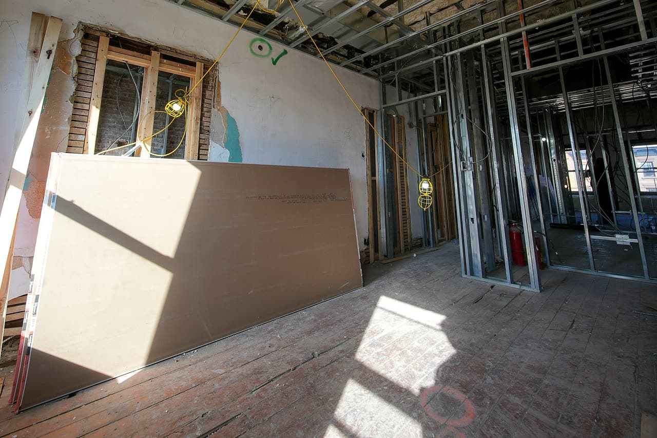 Sheets of drywall lean against a wall in a gutted room with new steel stud framing, spray-foam insulation overhead and temporary lights.
