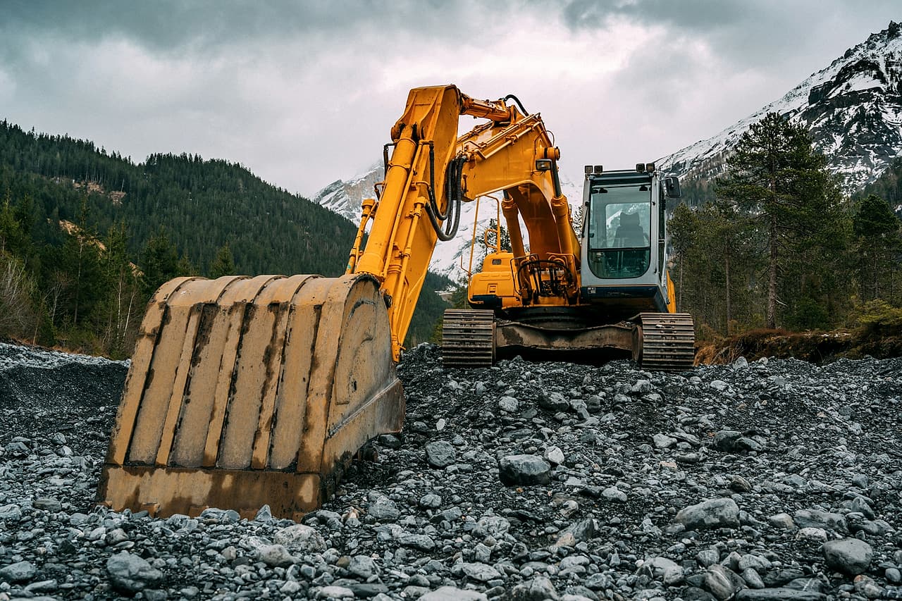 A yellow excavator sits idle on a gravel bed with its bucket lowered and the cab empty, forest and snowy mountains behind.