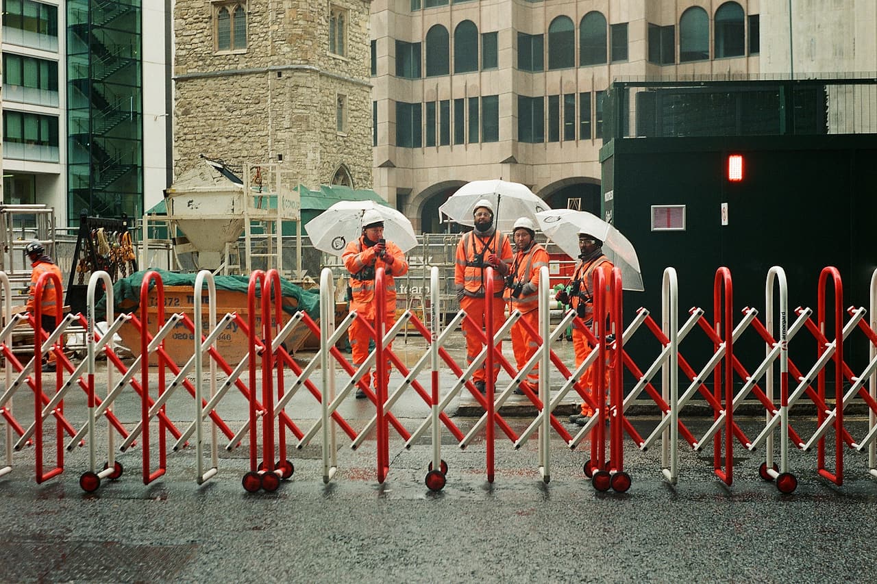 Construction workers in orange high-visibility gear and hard hats wait under umbrellas behind a red and white barrier on a wet city street.