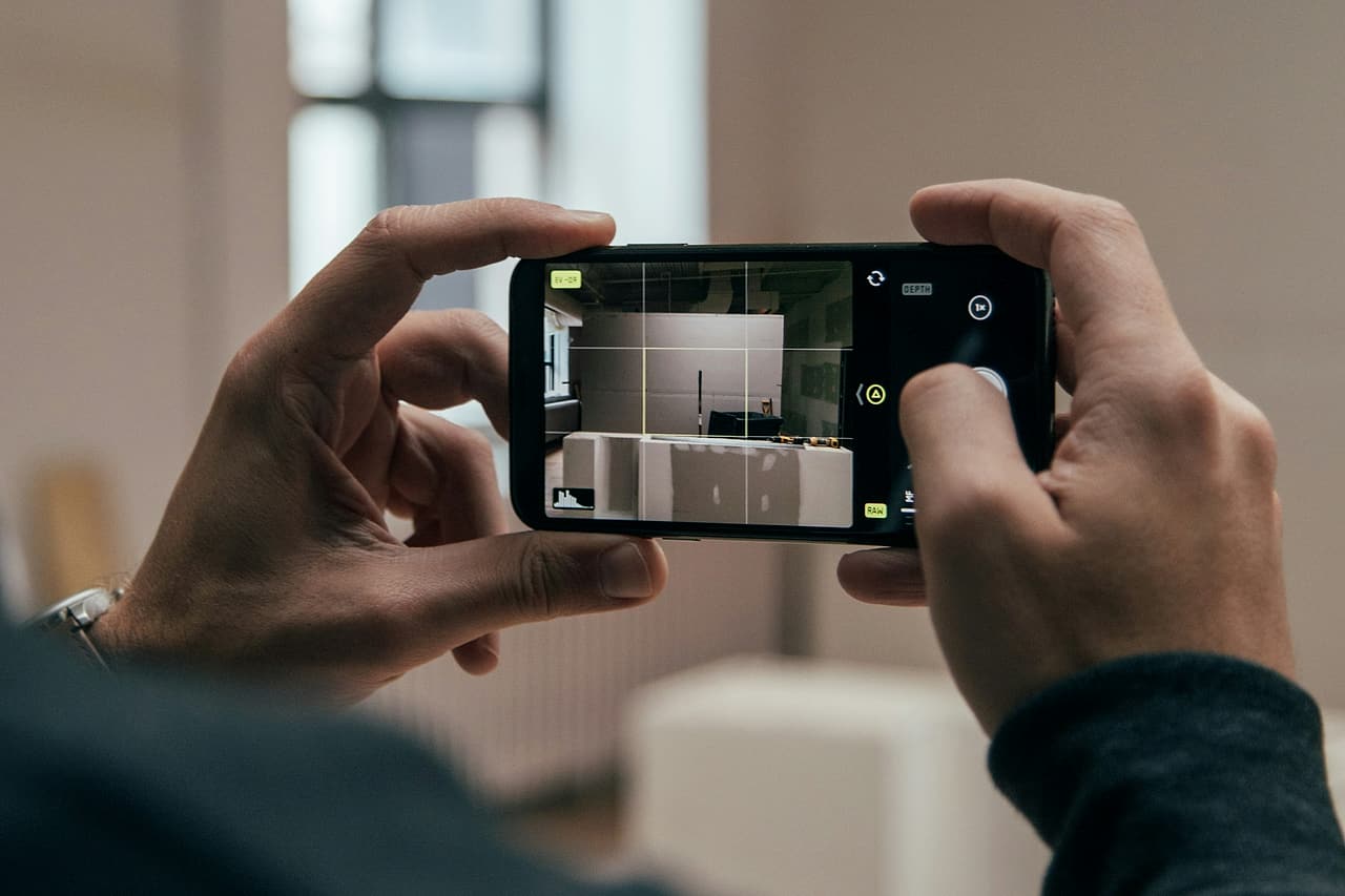 Seen over a shoulder, two hands hold a phone sideways with its camera set to RAW, framing a bare white interior room.