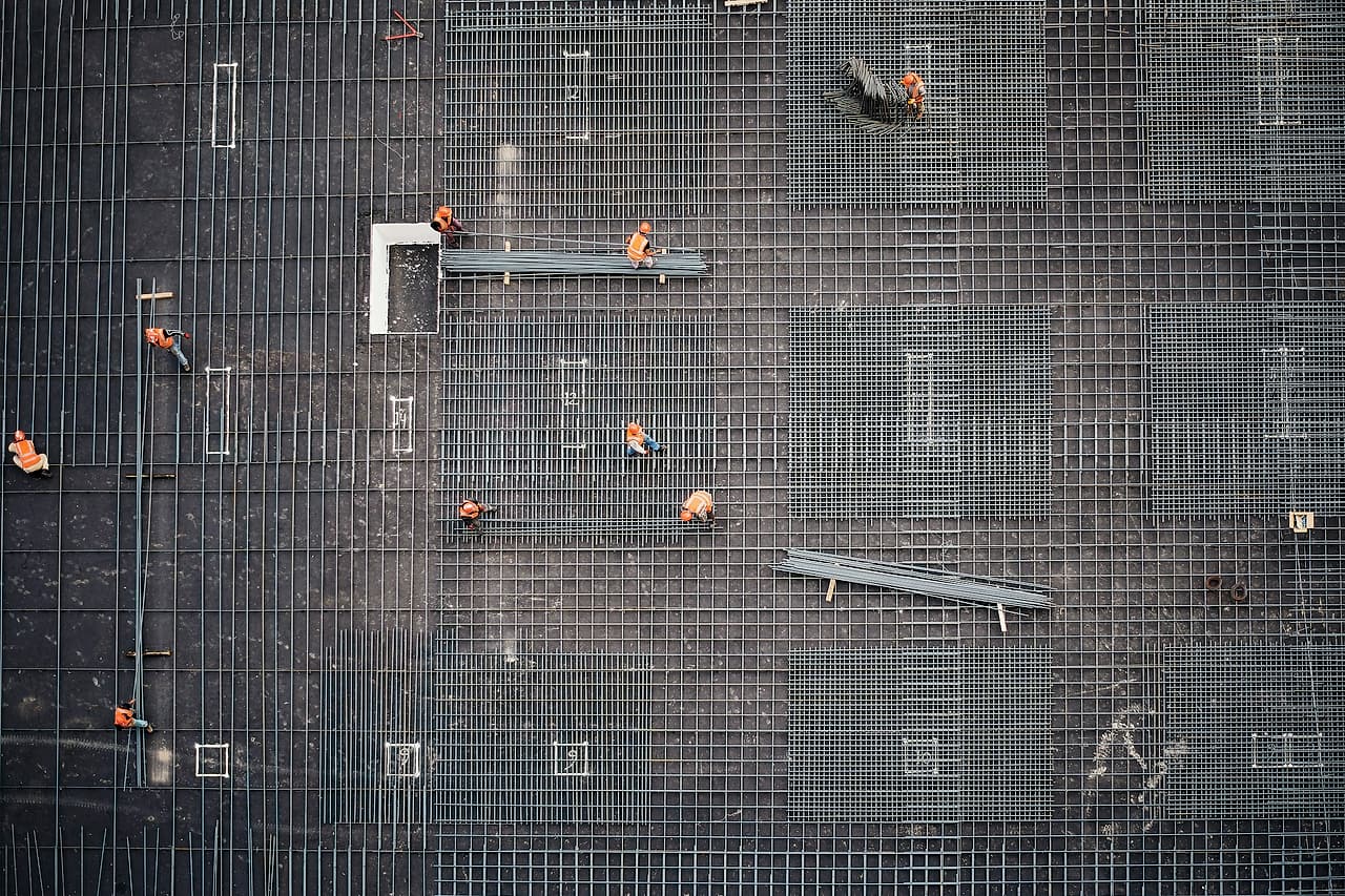 Seen from above, workers in orange vests and hard hats place and tie rebar across a large formed deck.
