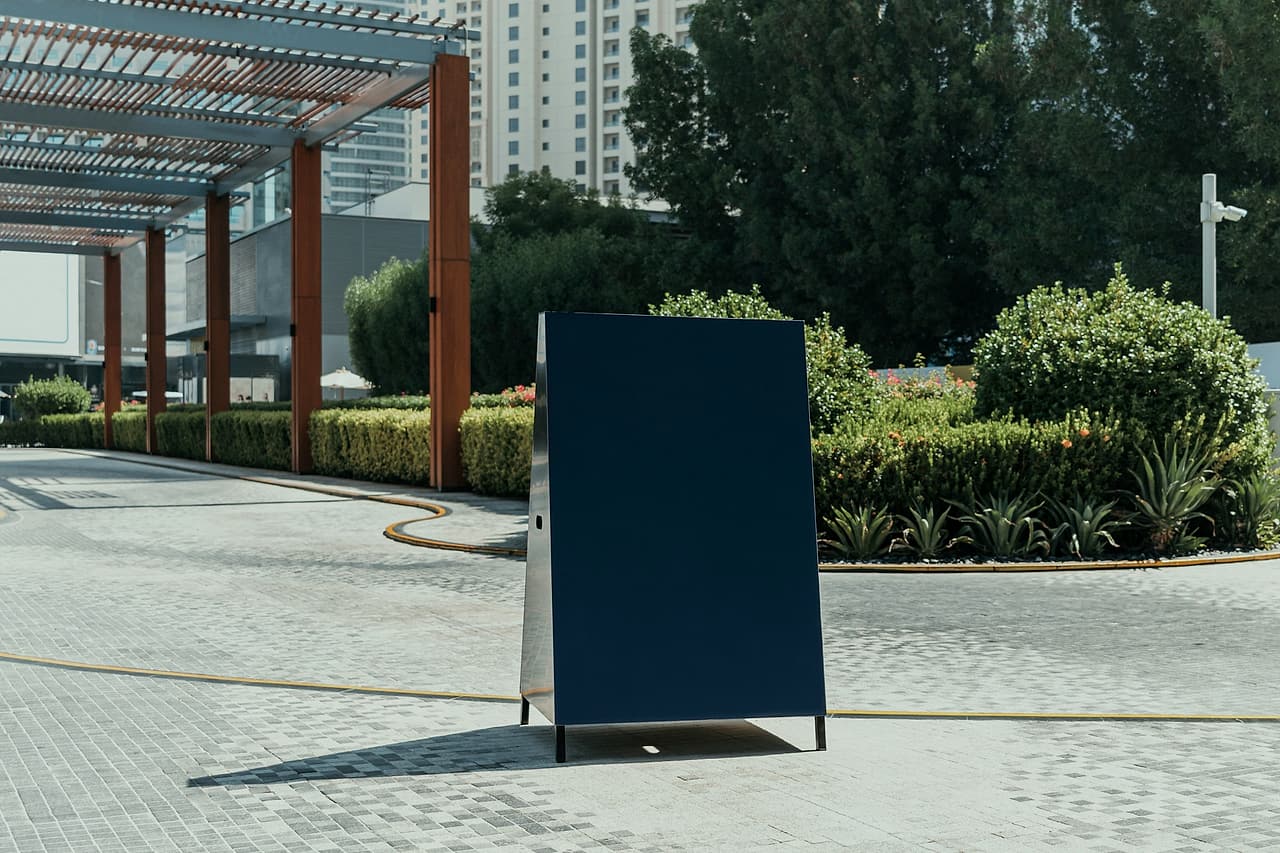 A blank freestanding notice board stands beside a paved pedestrian path and planted beds.