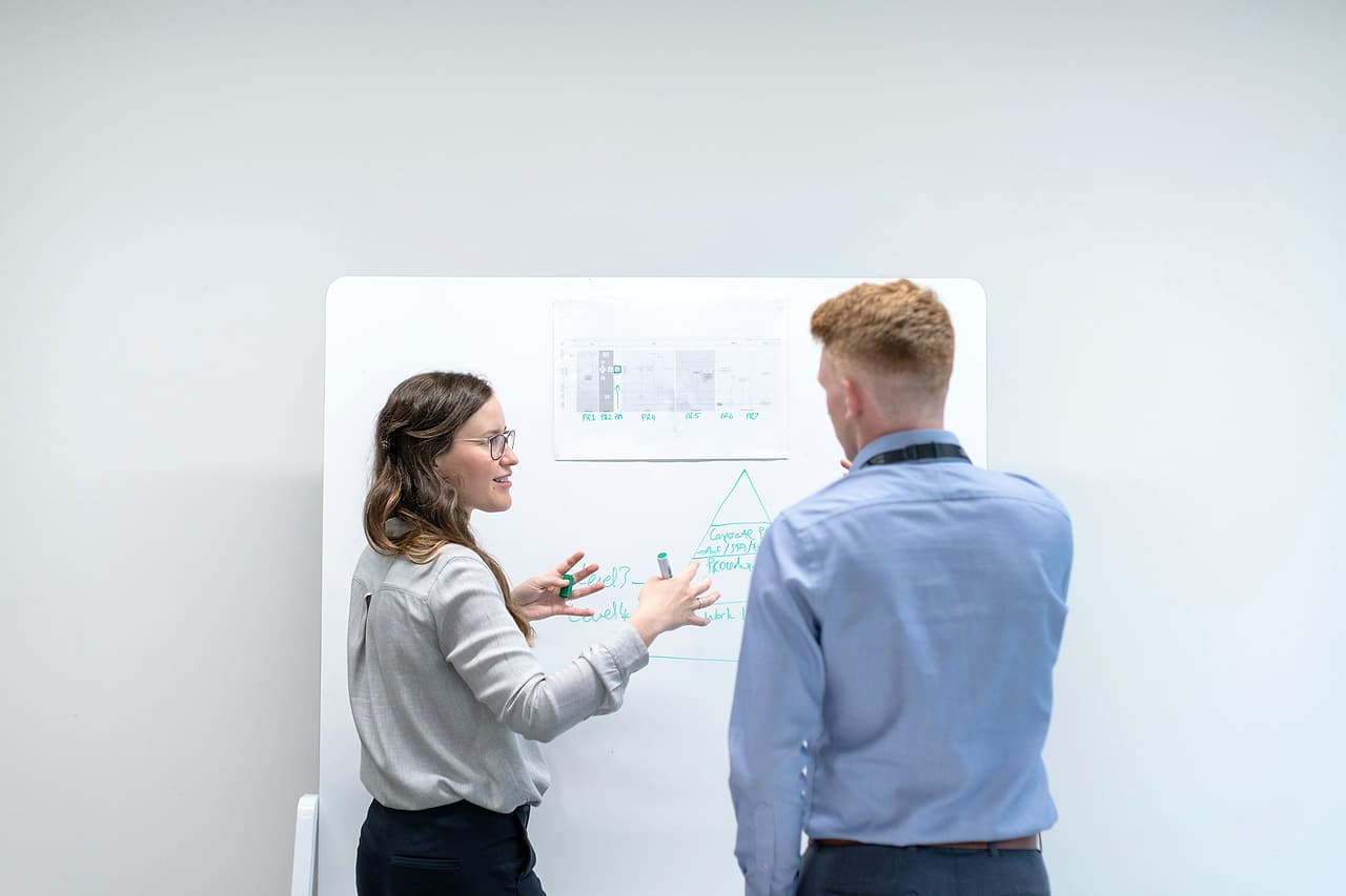 Two colleagues talk at a whiteboard with a printed chart pinned above their notes.