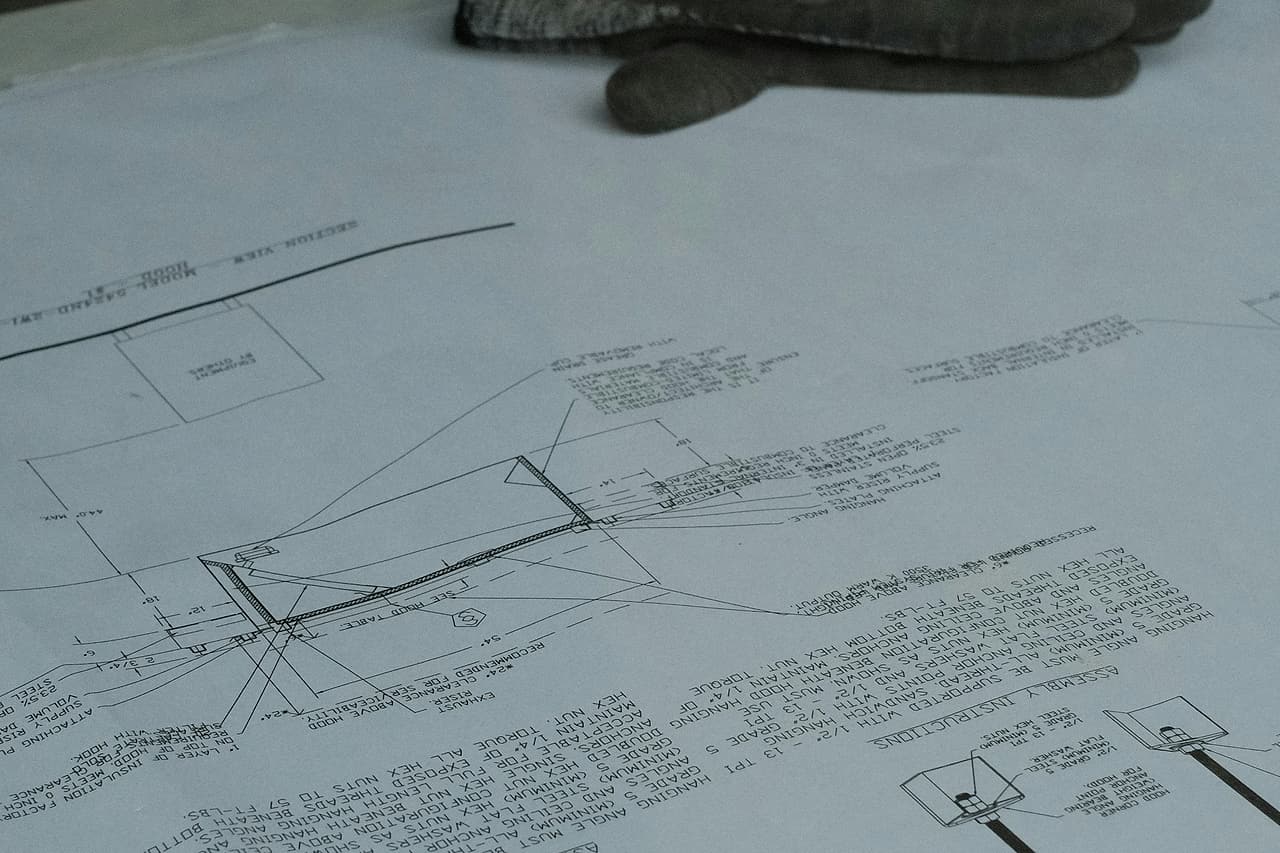 A shop drawing with assembly notes and details lies on a site table, a worn work glove resting on it, plywood stacked behind.