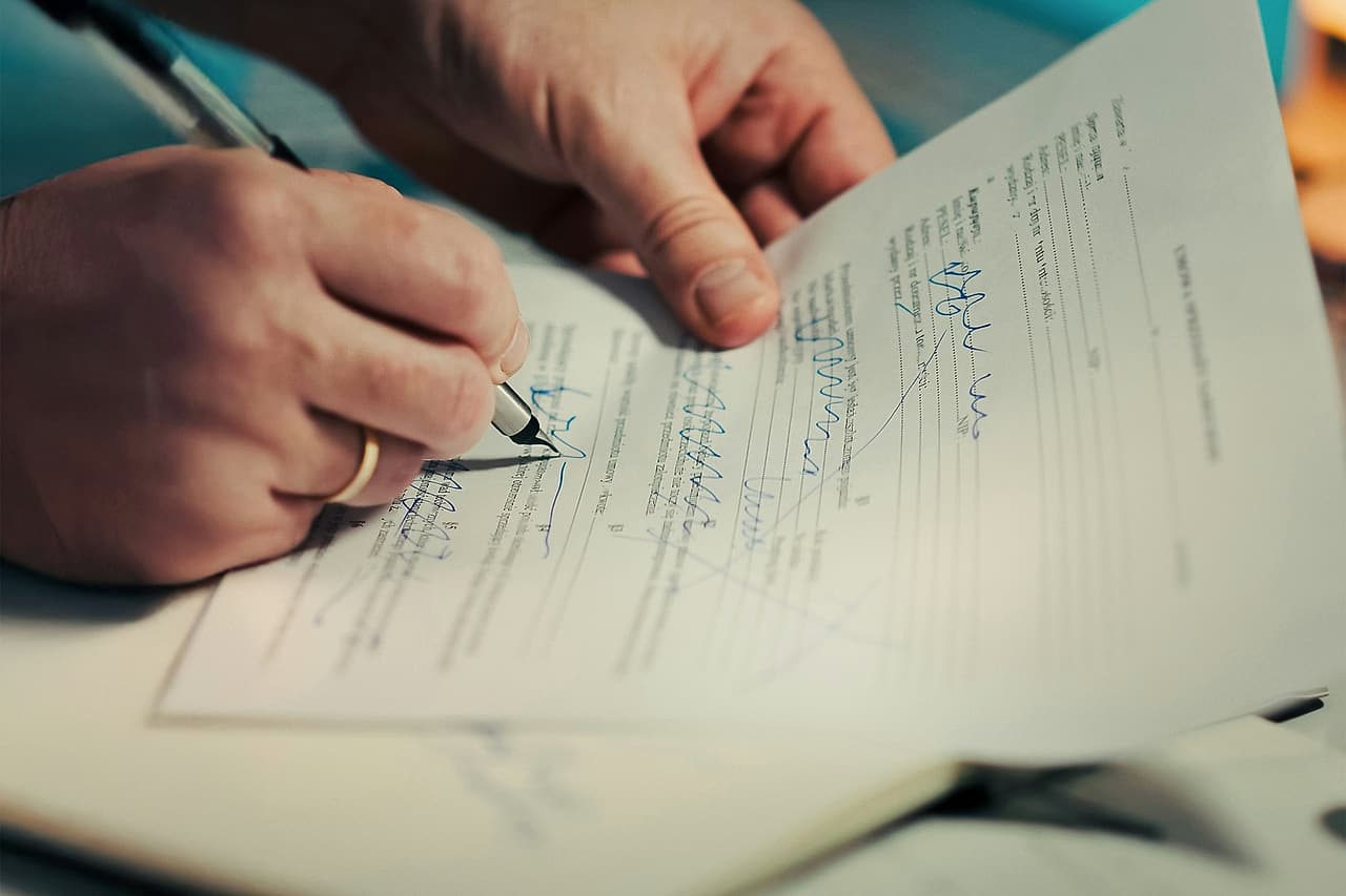 Two hands mark a printed form with a pen, notes in blue ink over its lines, at a desk with a laptop behind.