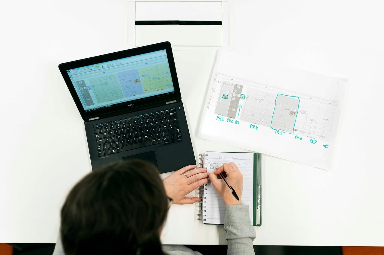 Seen from above, a person notes items in a spiral notebook beside a laptop showing a spreadsheet and a printed diagram marked in blue.
