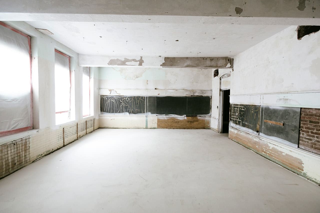 An empty institutional room stripped for renovation, with plastic over the windows, patched walls and old chalkboards.