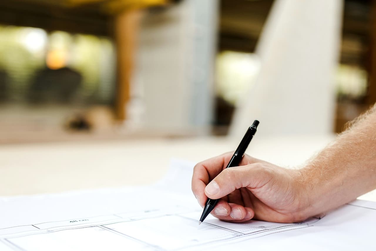 A hand marks a printed building plan with a black pencil.