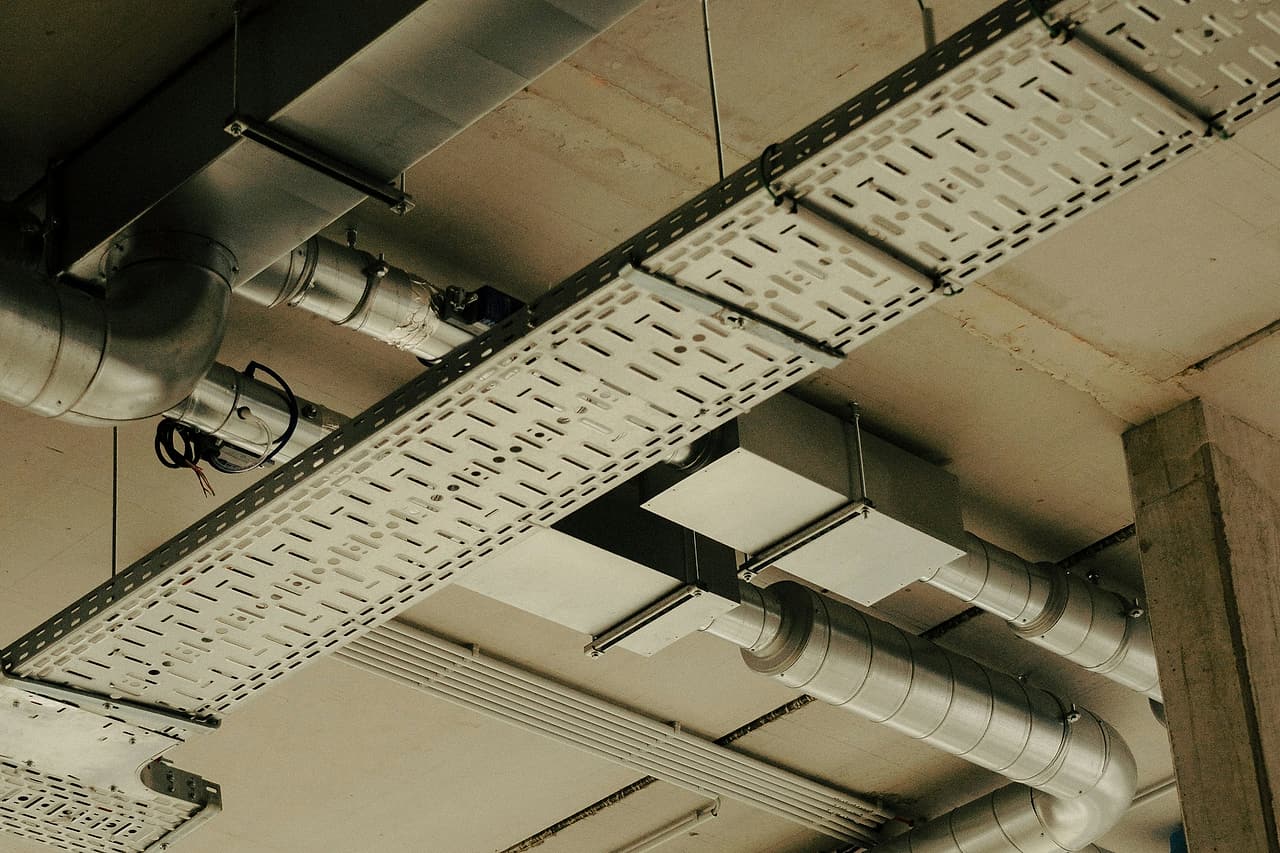 A perforated cable tray crosses beneath spiral ductwork and duct boxes on a concrete ceiling, next to a concrete column.