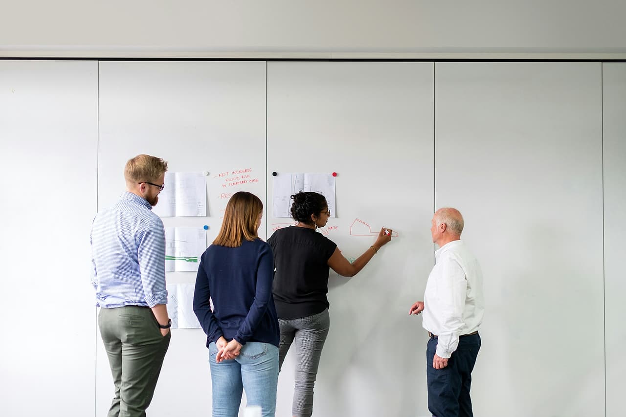 Four colleagues stand at a whiteboard wall with drawings pinned up while one writes notes in red marker.