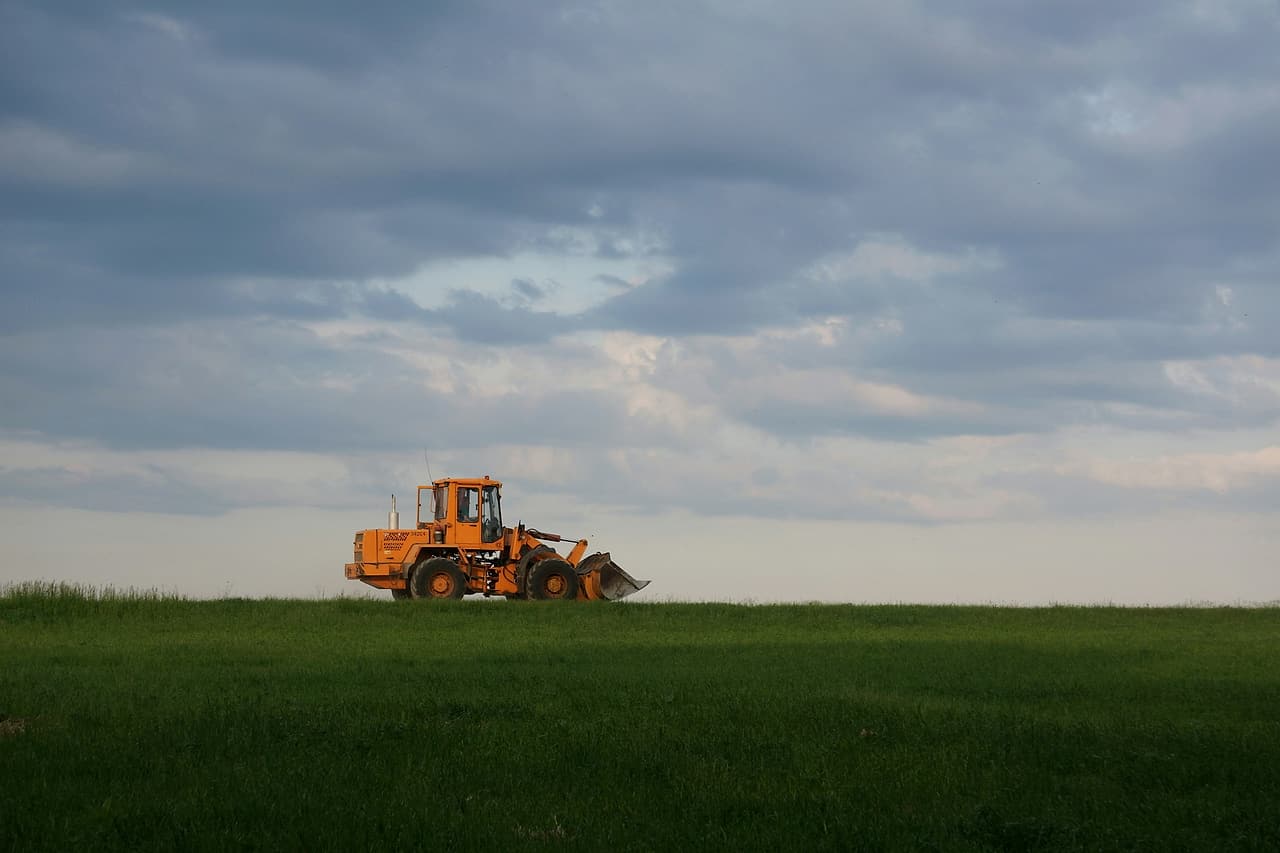 A lone wheel loader stands beyond a green field beneath a cloudy sky.