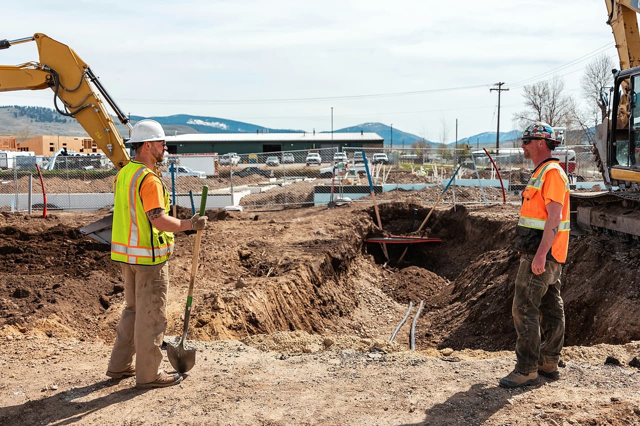 Two workers in hard hats and high-visibility vests talk across a sloped excavation, an excavator arm above them.