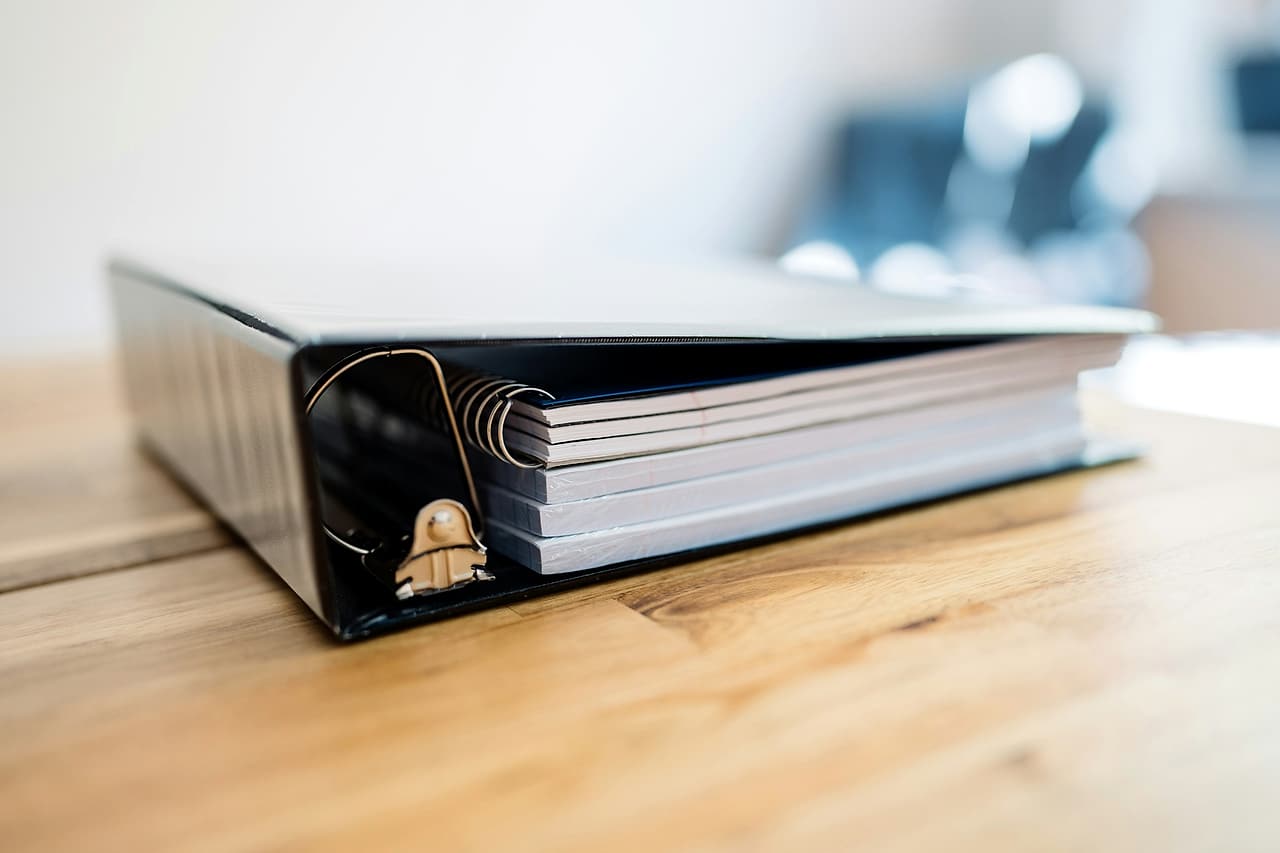 A thick black ring binder packed with bound documents lies on a wooden desk.