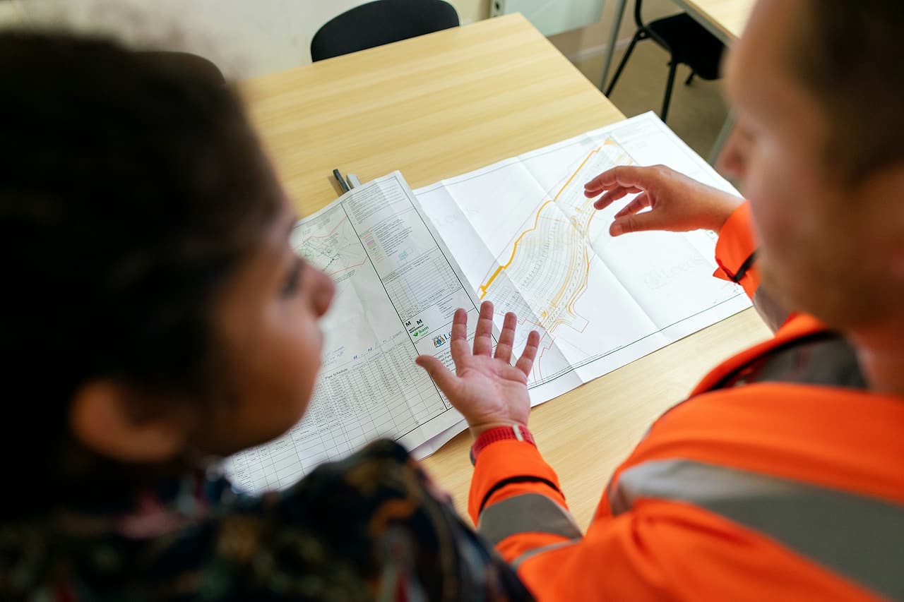 Two people, one in an orange high-visibility jacket, go over a site drawing together at a table.
