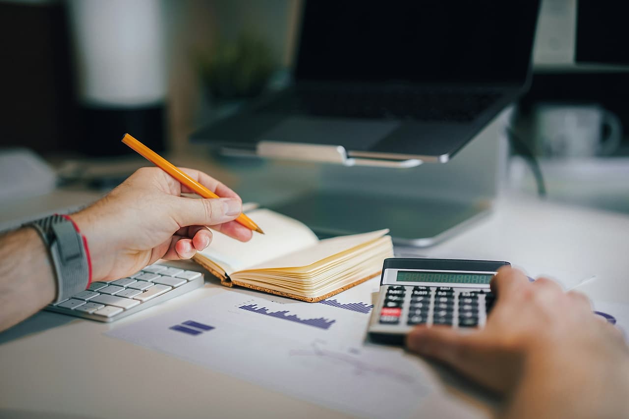 First-person view of a desk: one hand writes in a notebook with a pencil, the other works a calculator over printed charts, a laptop beyond.