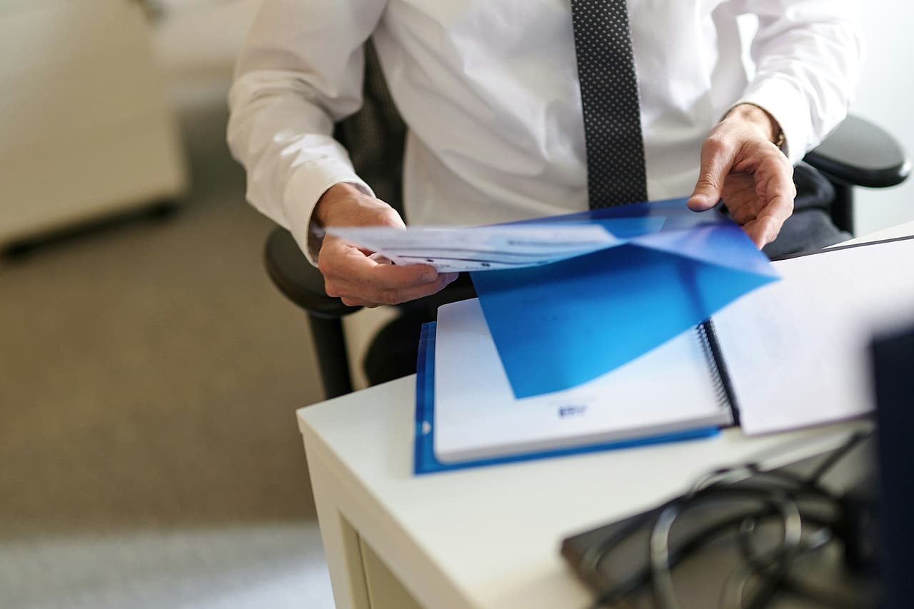 A person in a white shirt and tie slides printed pages into a blue folder at a white desk.