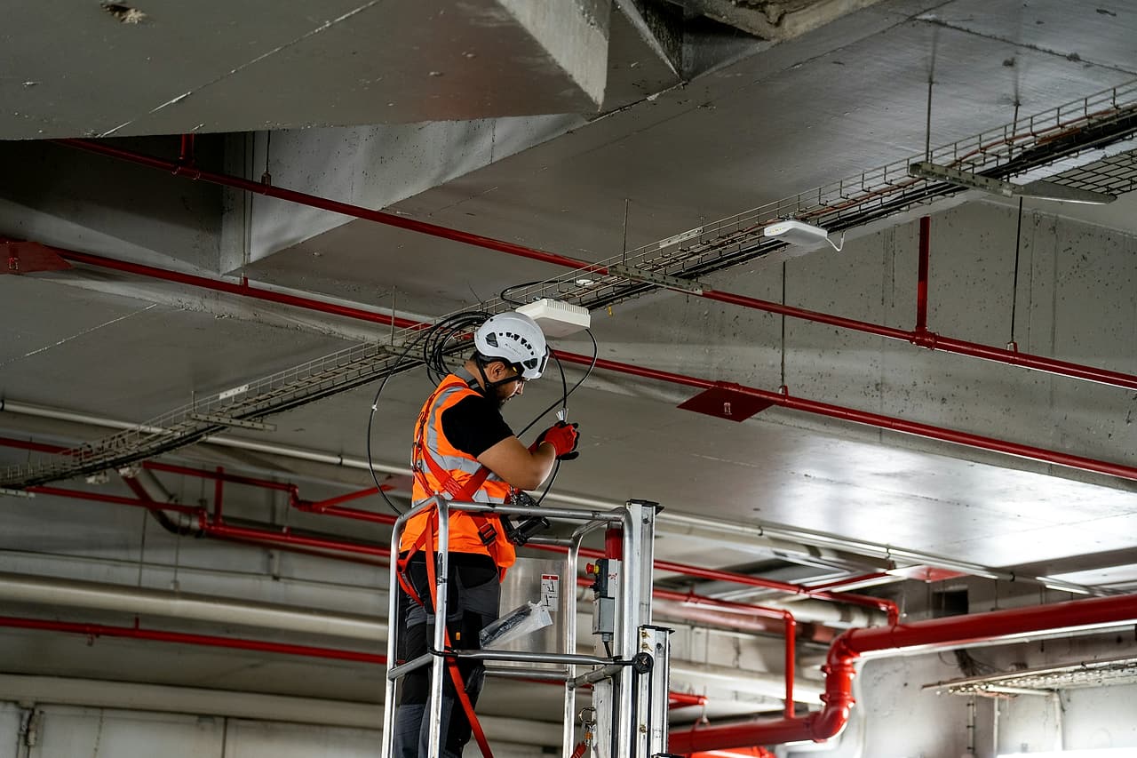 A technician in a helmet, harness and high-visibility vest works from a lift platform among cable trays and red sprinkler pipes.