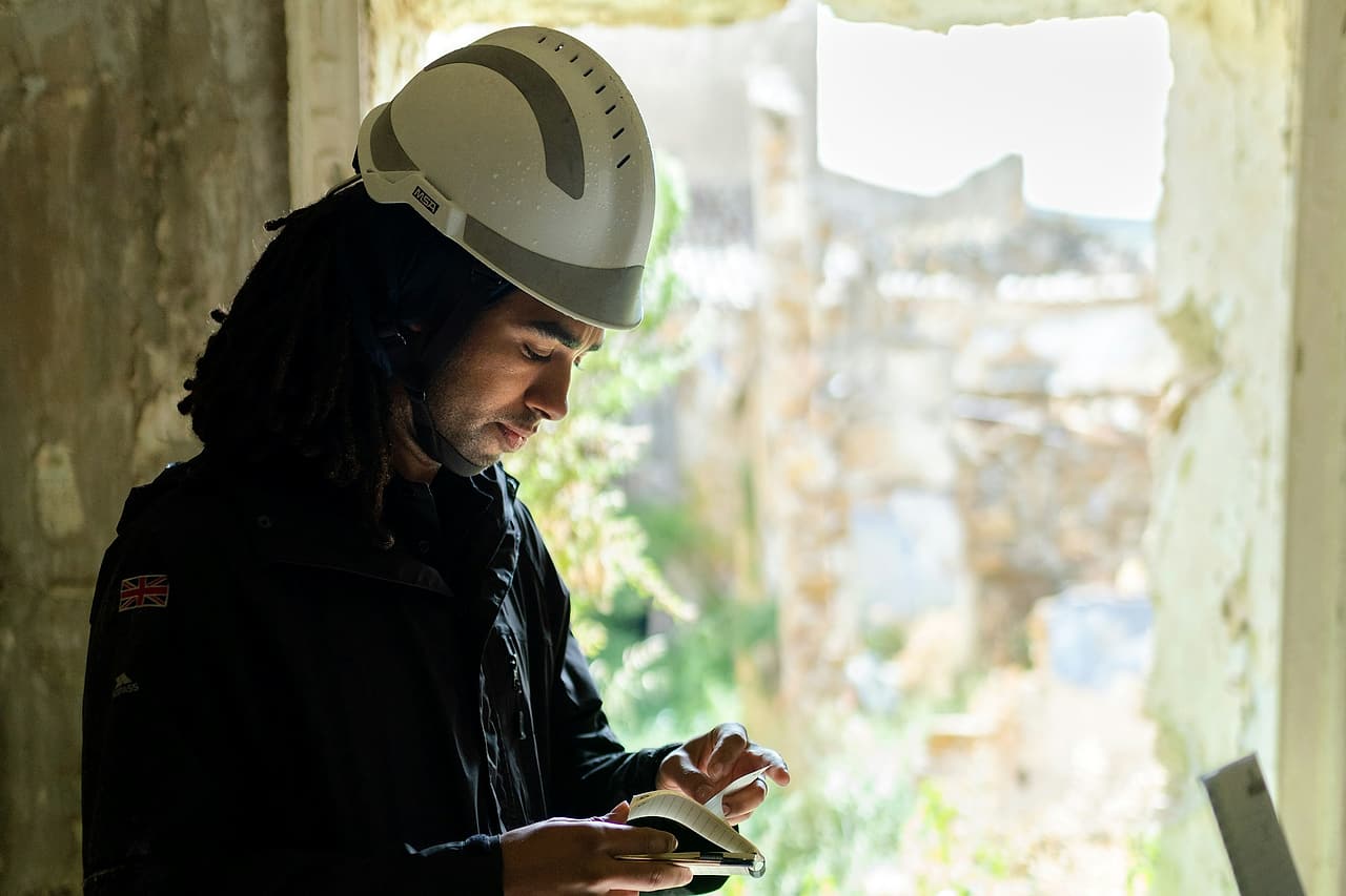 A person in a white hard hat flips through a pocket notebook, pen in hand, inside an old stone building.